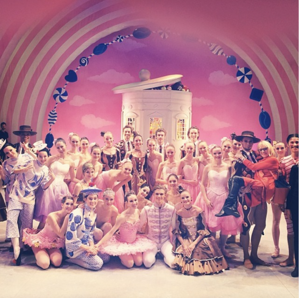 A large group of ballet dancers stands onstage in an array of costumes, everyone smiling at the camera.